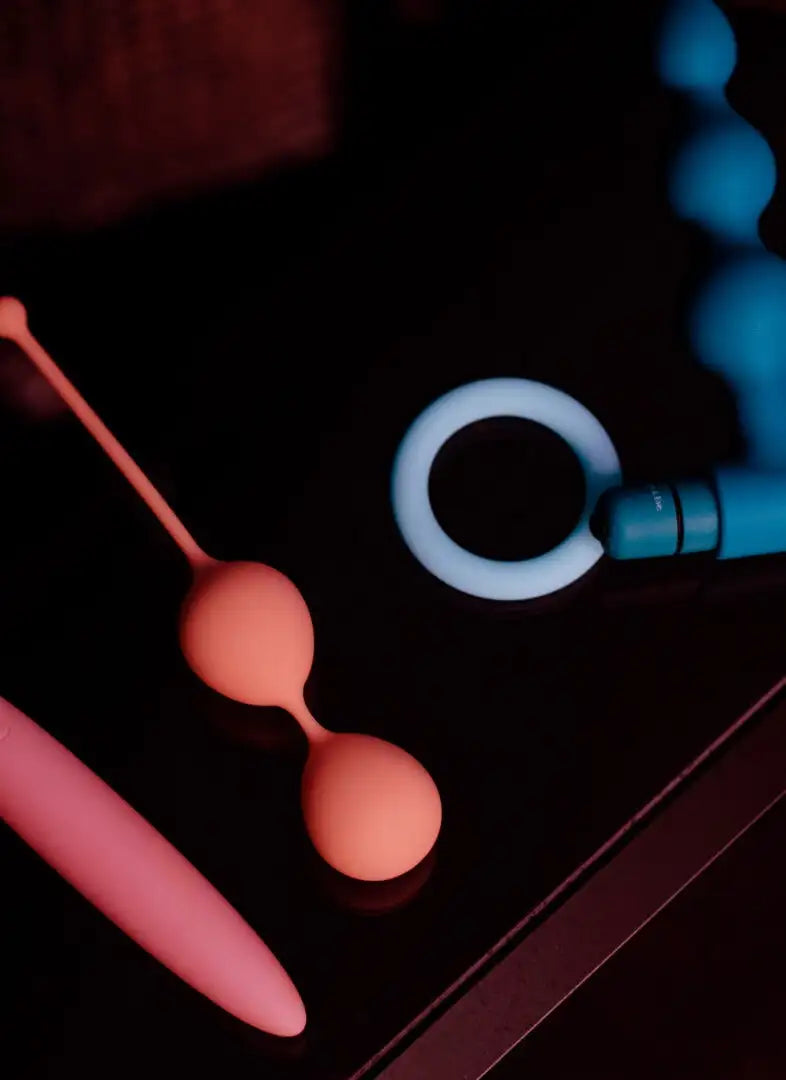 A peach-colored silicone double-balls set with a slender handle, resting beside other vibrators on a dark surface.slider_item_zW8Fik