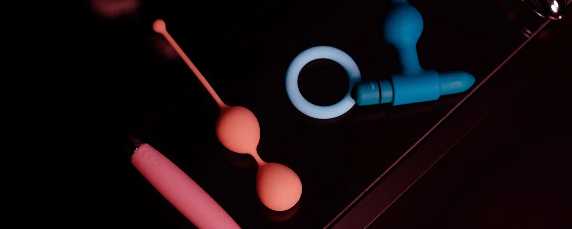 A peach-colored silicone double-balls set with a slender handle, resting beside other vibrators on a dark surface.slider_item_zW8Fik